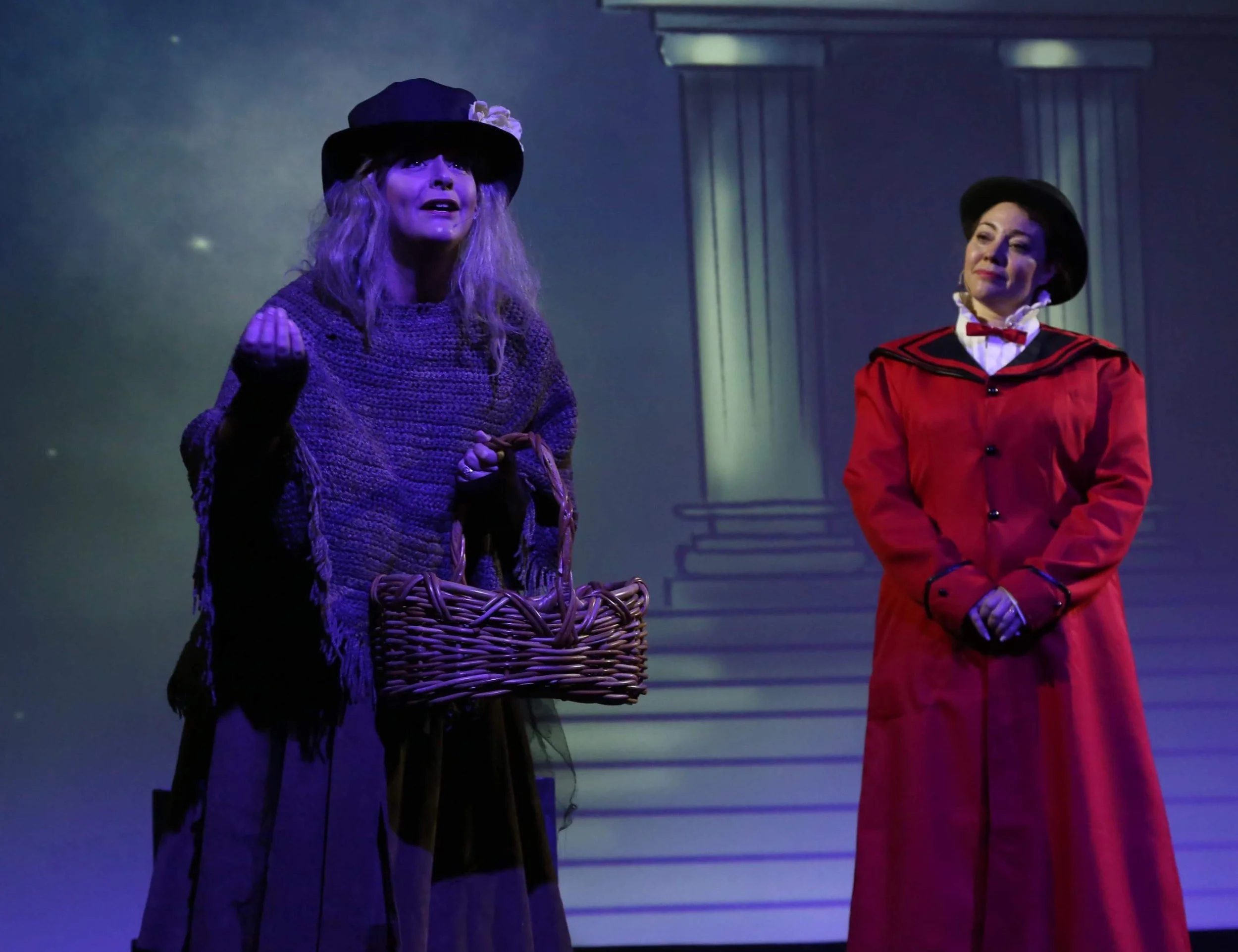 Two women stand on a stage with columns in the background, one woman wearing a purple outfit and a black hat with a flower, holding a wicker basket, and the other woman wearing a red outfit and a black hat, with a smile.