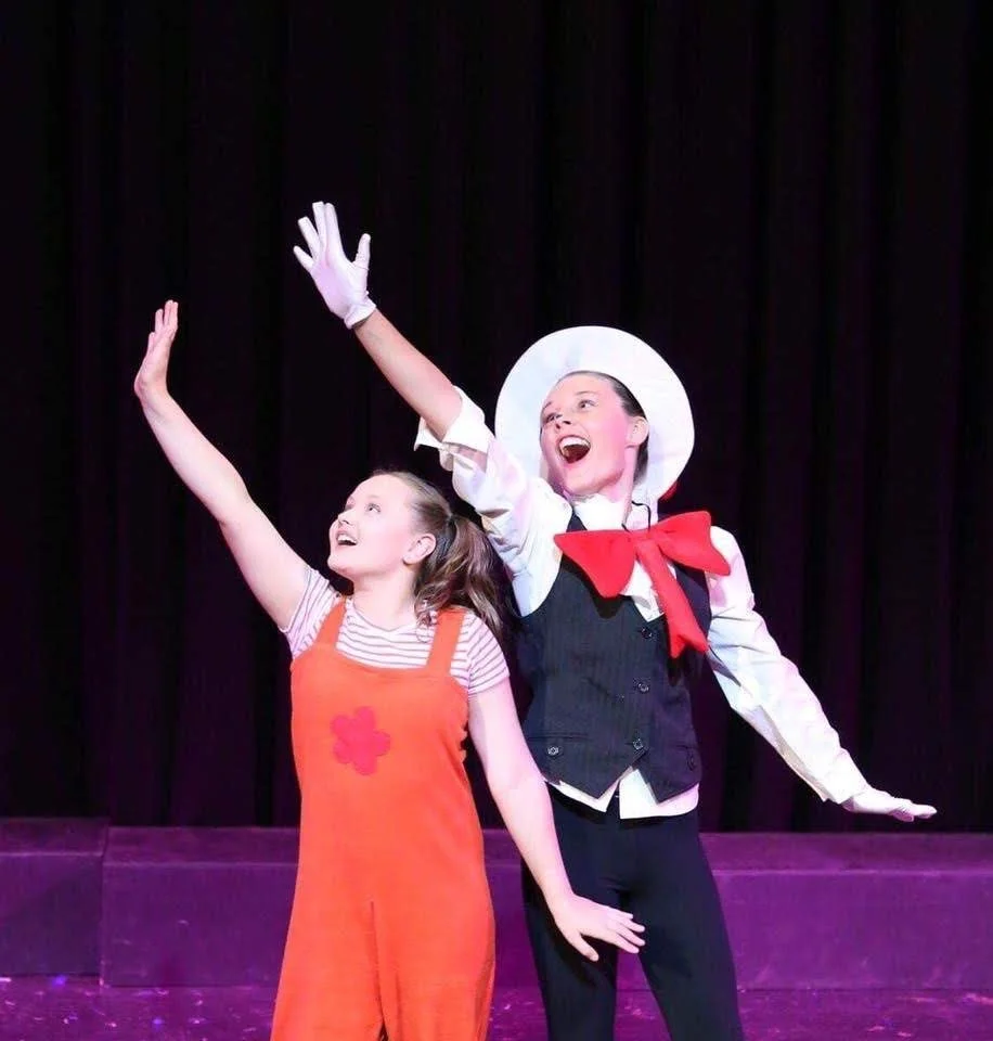 Two girls performing on stage, one in an orange costume with a pink star, the other in a white hat, black vest, and red scarf, both smiling and raising their arms.