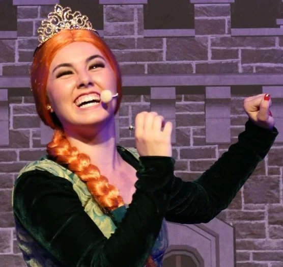 A woman dressed as a princess with a crown, orange braided hair, and a microphone headset, smiling and posing with her arm flexed in front of a brick wall.