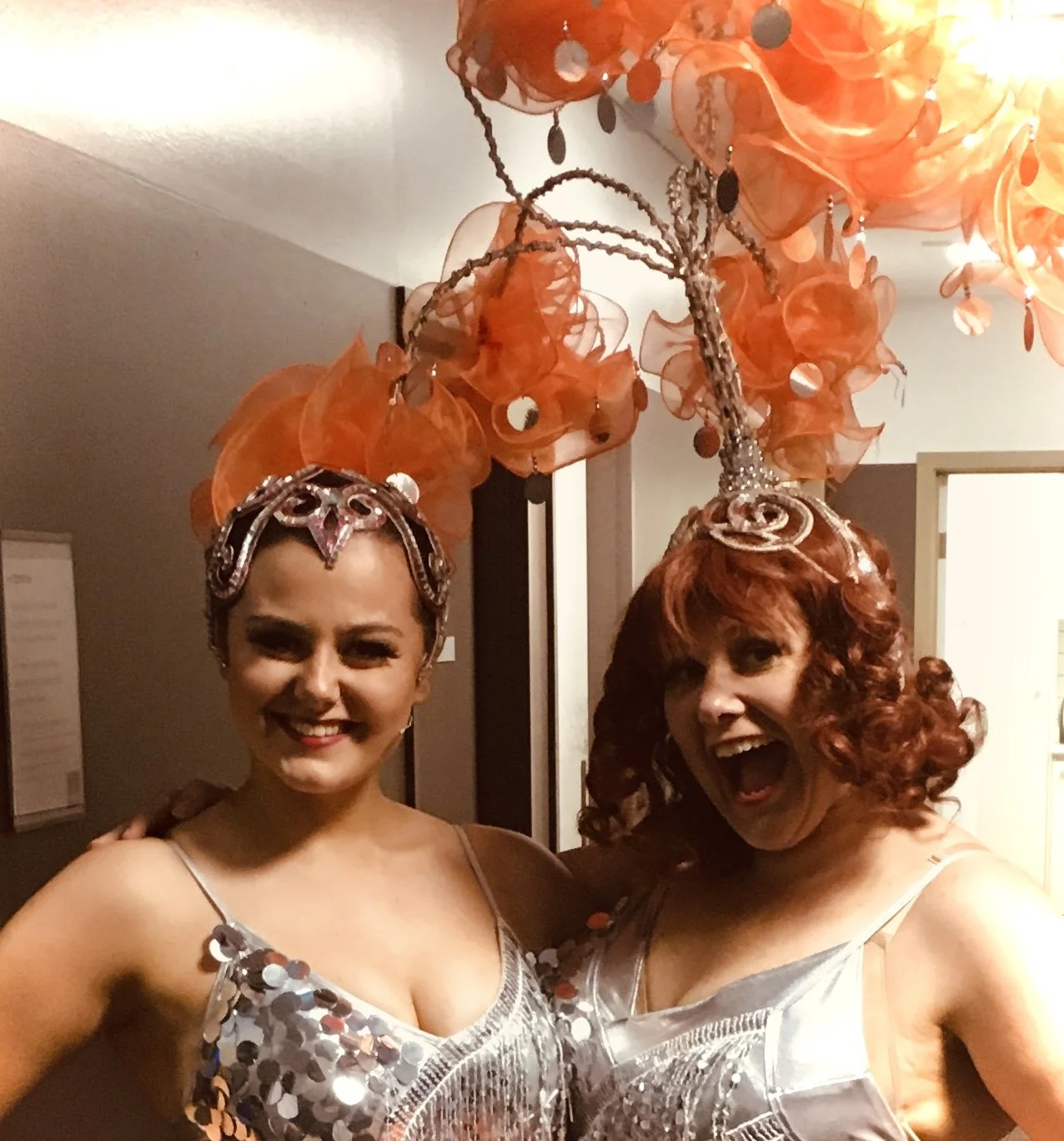 Two women in sequined outfits and elaborate headpieces, one with a playful wink and the other smiling widely, standing under an orange decorative chandelier.