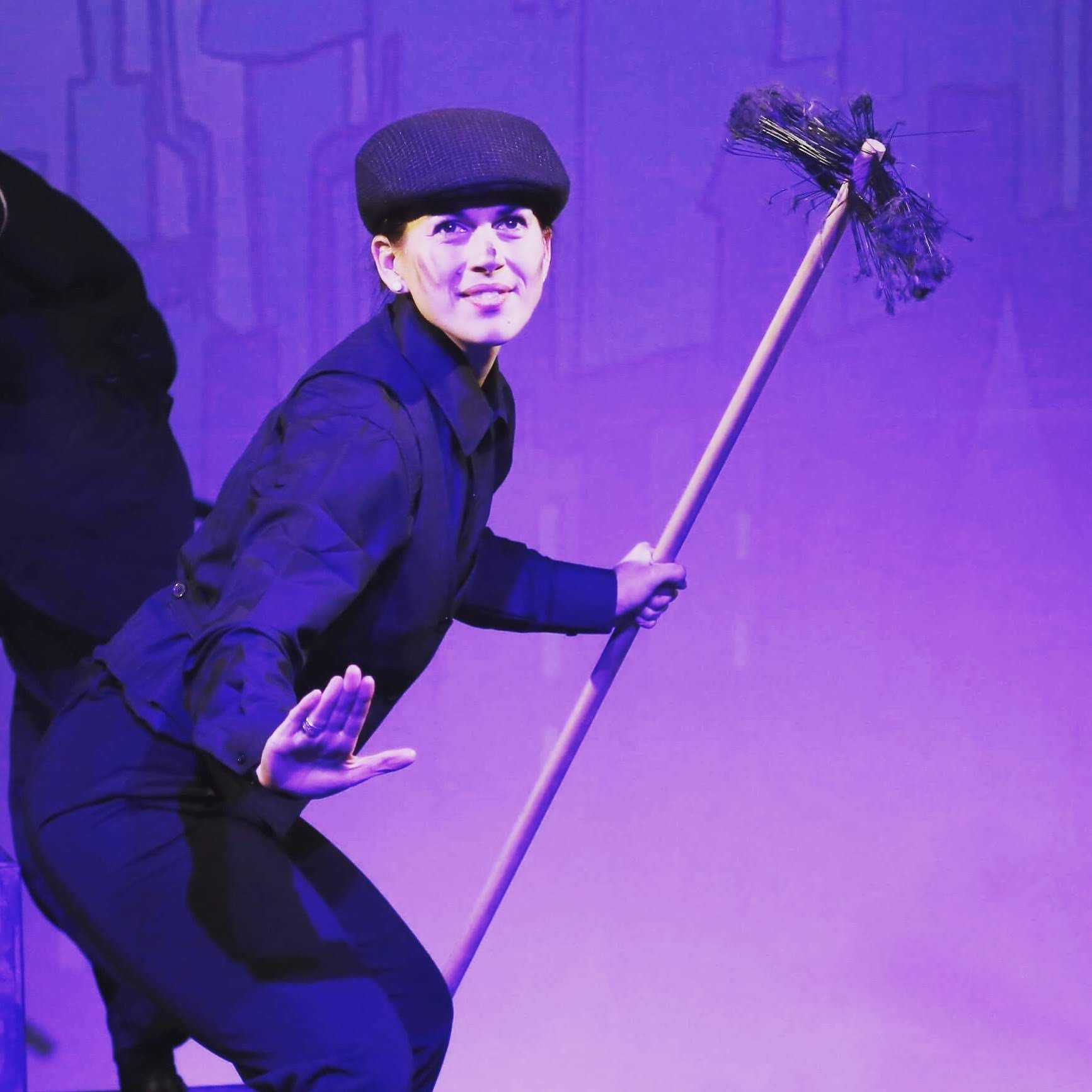 A woman in a black jacket and black hat holding a broomstick, with purple stage lighting in the background.