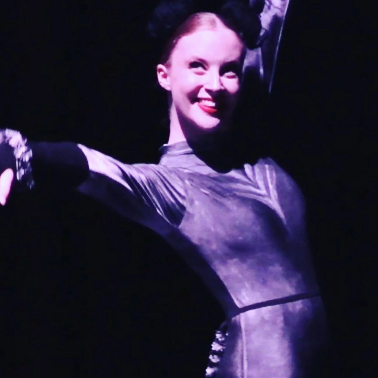 A person with a smile, dressed in a shiny outfit, on stage with dark background, wearing black cat ears headband.