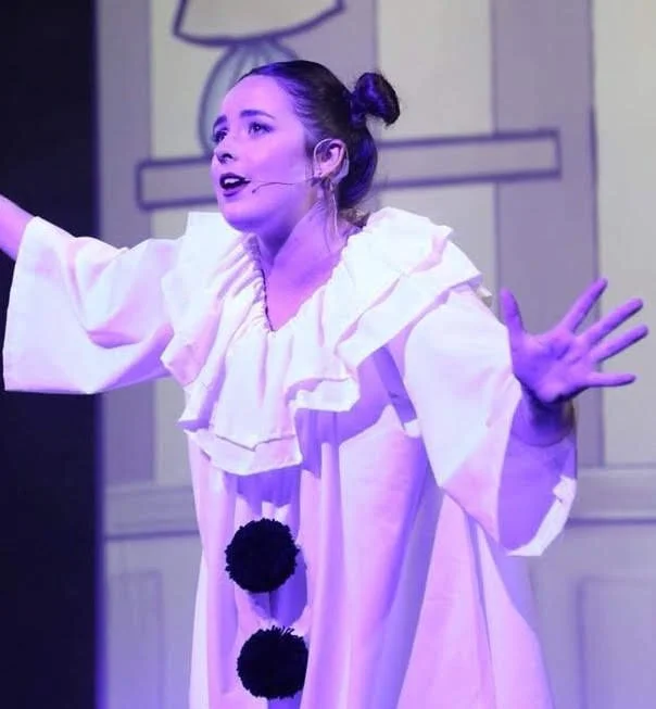 A woman performing on stage, wearing a clown costume with large pom-poms, and a headset microphone, gesturing with her hands.