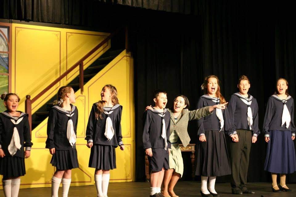 Group of children on stage dressed as sailors, with one woman in a gray coat, performing in a theatrical play.