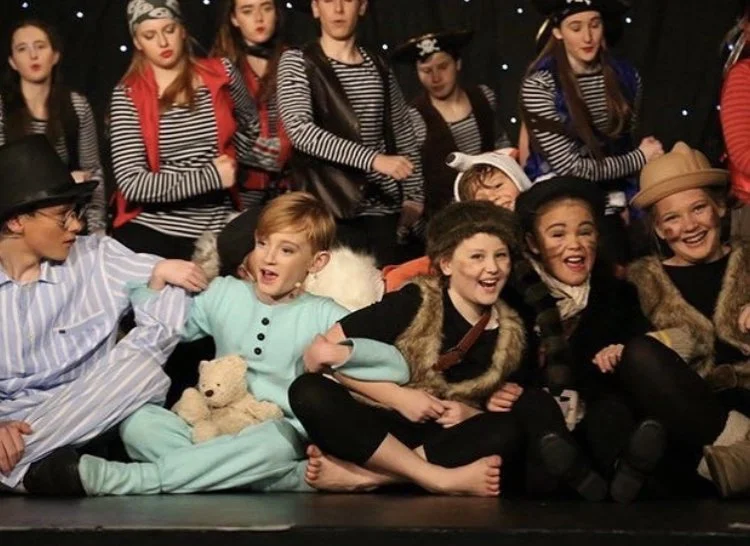 Children dressed in costumes, some in pirate outfits and others in pajamas, seated on a stage, in a  theater setting, during a performance or rehearsal.