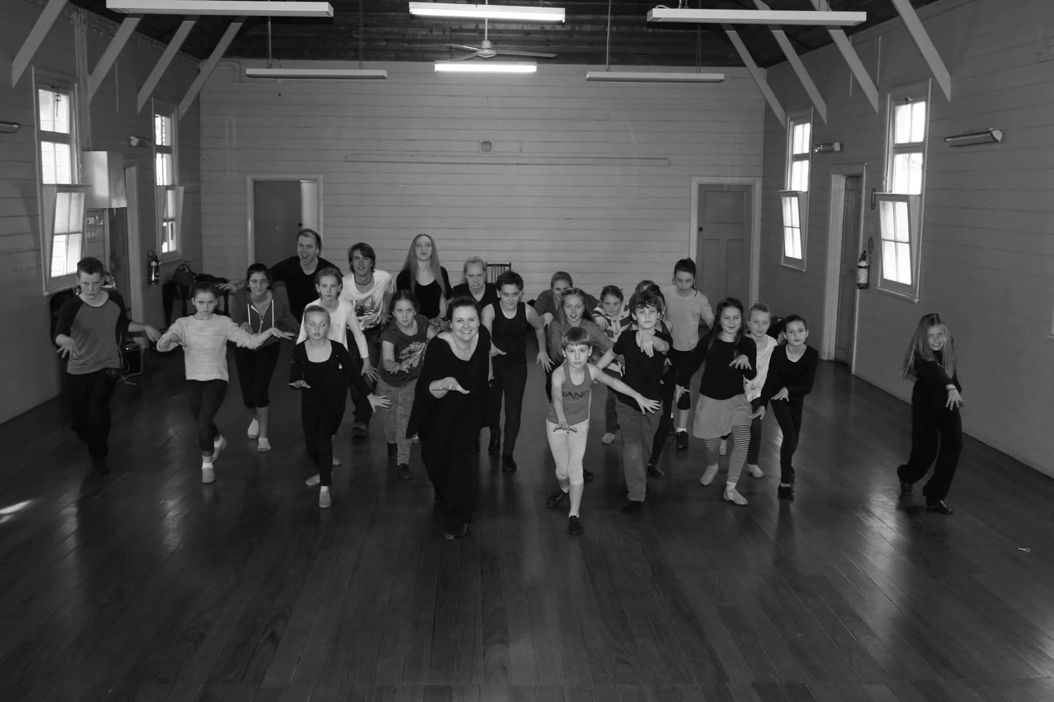 A group of children and adults practicing dance in a spacious wooden-floored room with high ceilings and large windows.