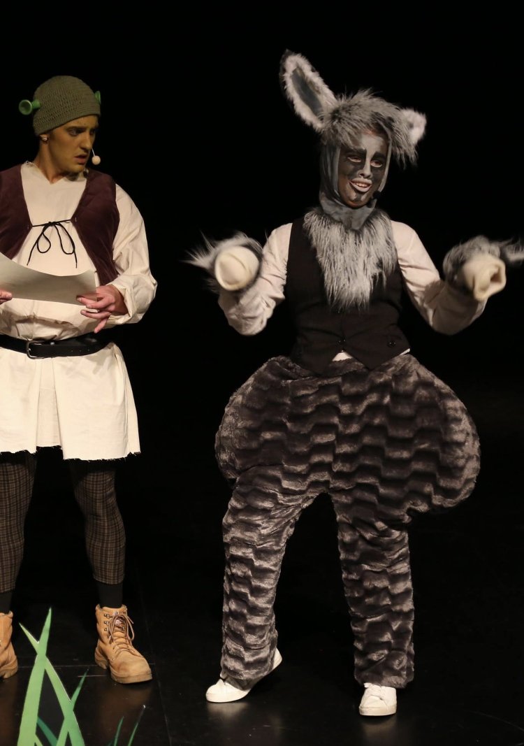 Two actors on stage in costume, one dressed as a gray and white donkey with large ears and fluffy tail, and the other in a costume with a knit hat and a brown vest. The donkey actor is smiling and making a gesture with gloved hands.