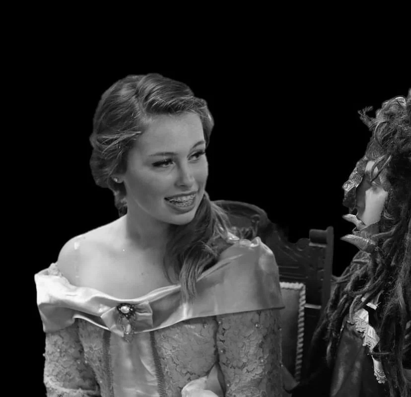 A black and white photo of two people talking. One woman has wavy hair, is smiling, and wearing an off-shoulder dress with lace. The other person has curly hair and glasses, and is dressed in dark clothing.
