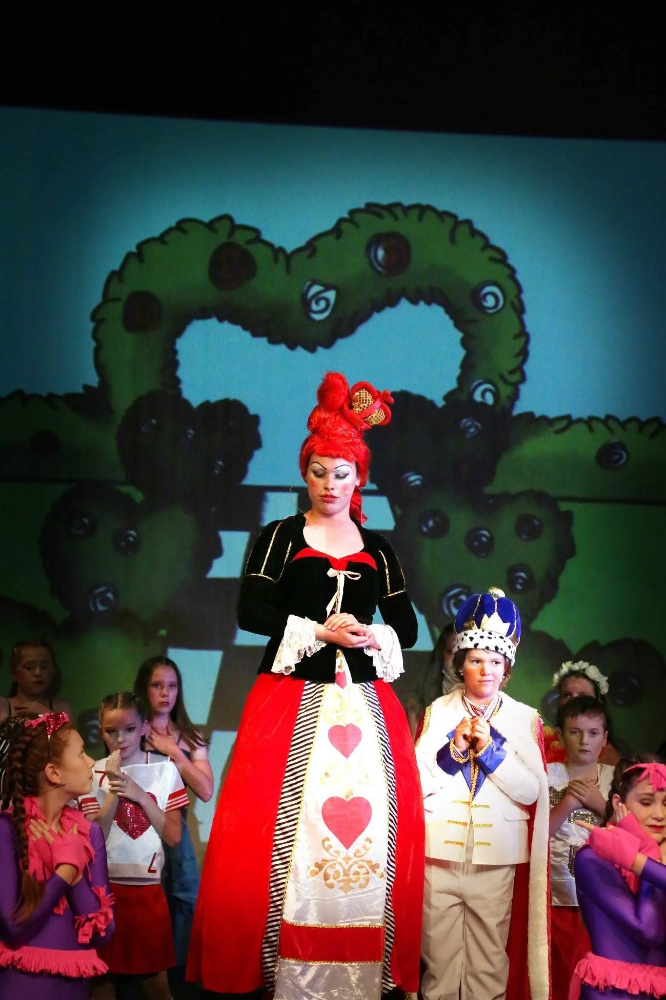 Children and adults in colorful costumes participating in a theatrical performance on stage, with a backdrop of a cartoonish green landscape with a large arch and trees.