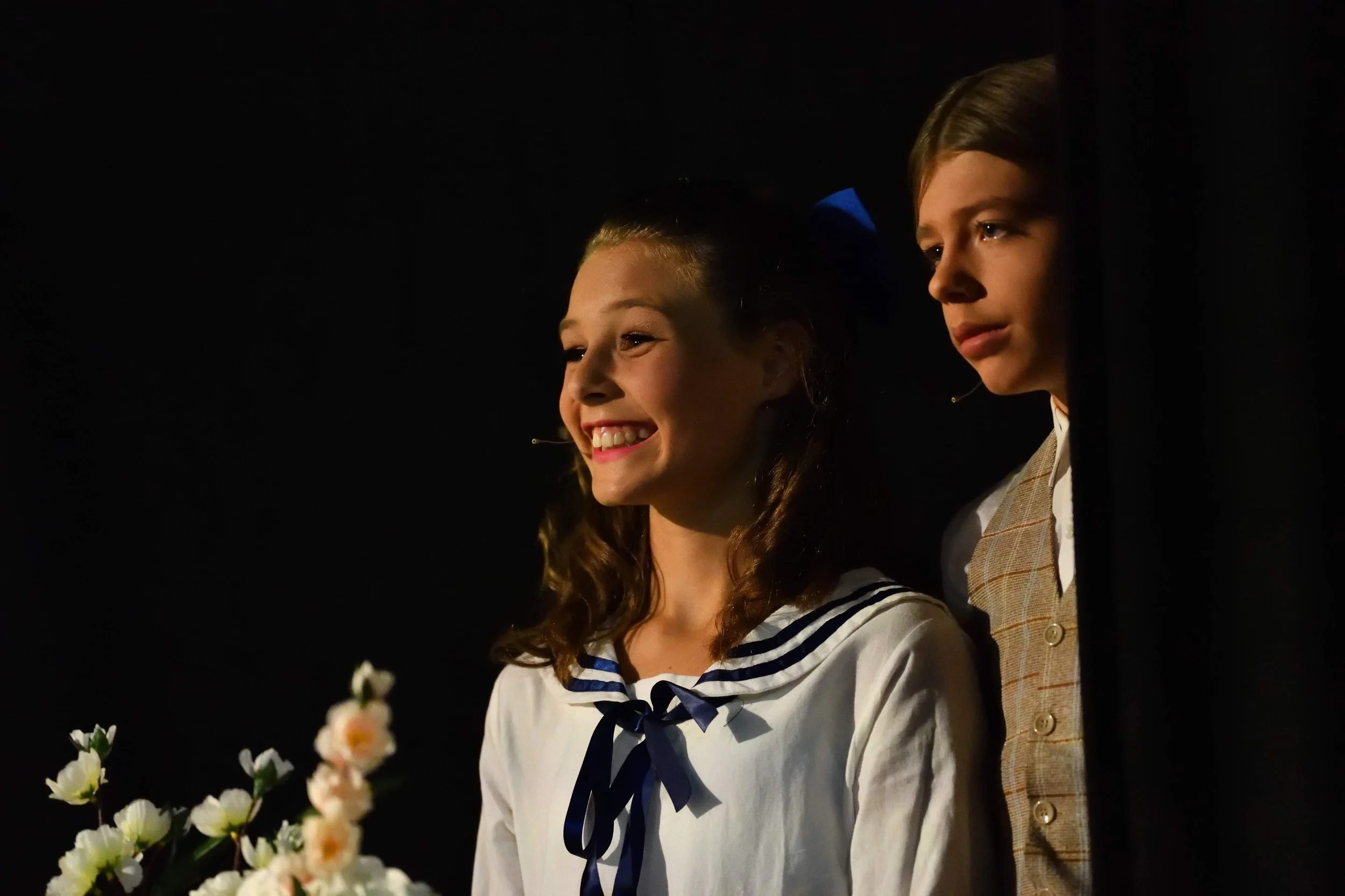 Two children, a girl with curly hair and a boy with straight hair, both wearing school uniforms, are looking at something off-camera in a dark setting. The girl is smiling, and the boy appears serious. There are flowers in the foreground.
