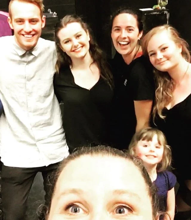 Group of five people smiling for a selfie indoors, with one person at the bottom taking the photo. Four people are standing behind her, and a young girl is in front of them. Everyone appears happy and relaxed.