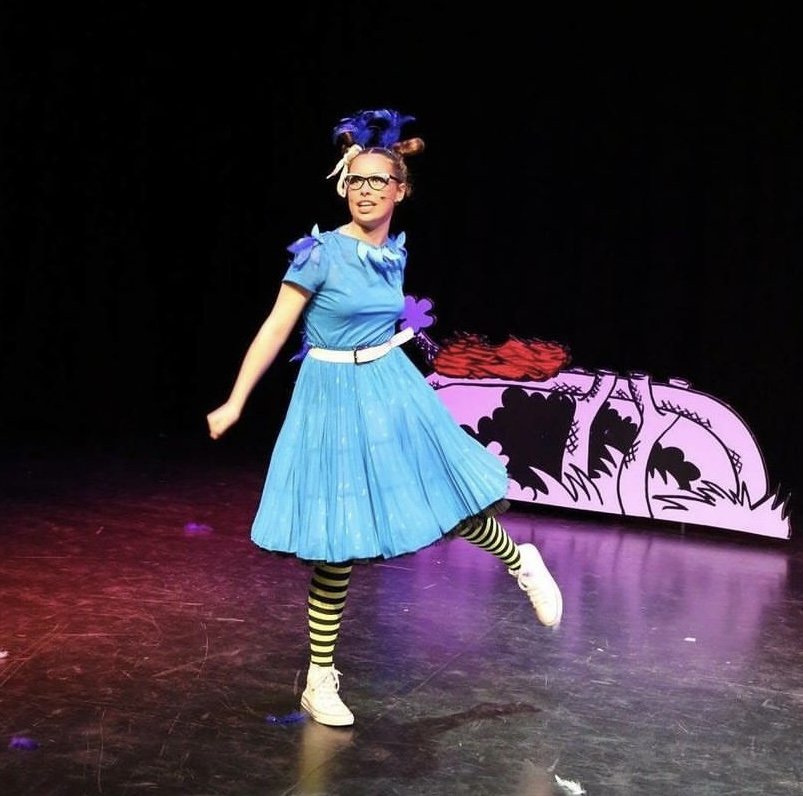 Performer in colorful costume on stage with black background and pink and purple props.