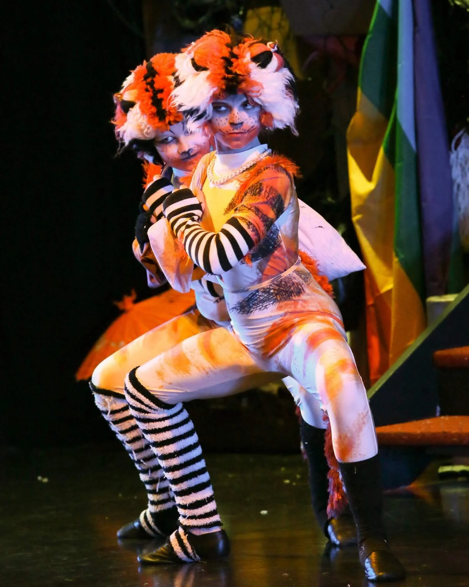 Two children dressed in cat costumes performing on stage, with black background and some stage equipment visible.