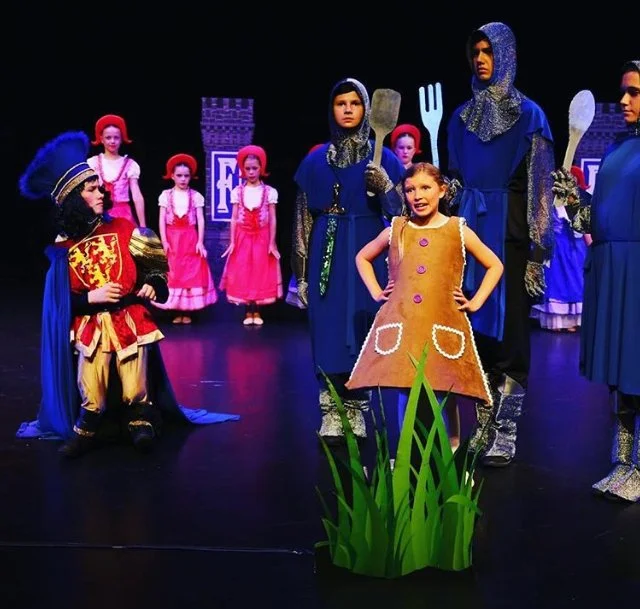 Children performing a play on stage dressed as medieval characters and fairytale figures, with a castle backdrop and props resembling grass.