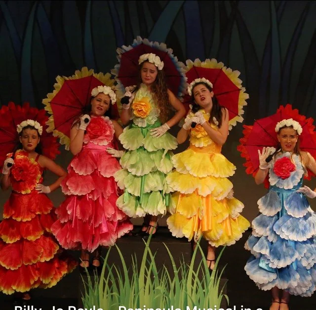 Four women in colorful layered dresses and floral headpieces holding parasols perform on stage with a painted backdrop of tall grass.