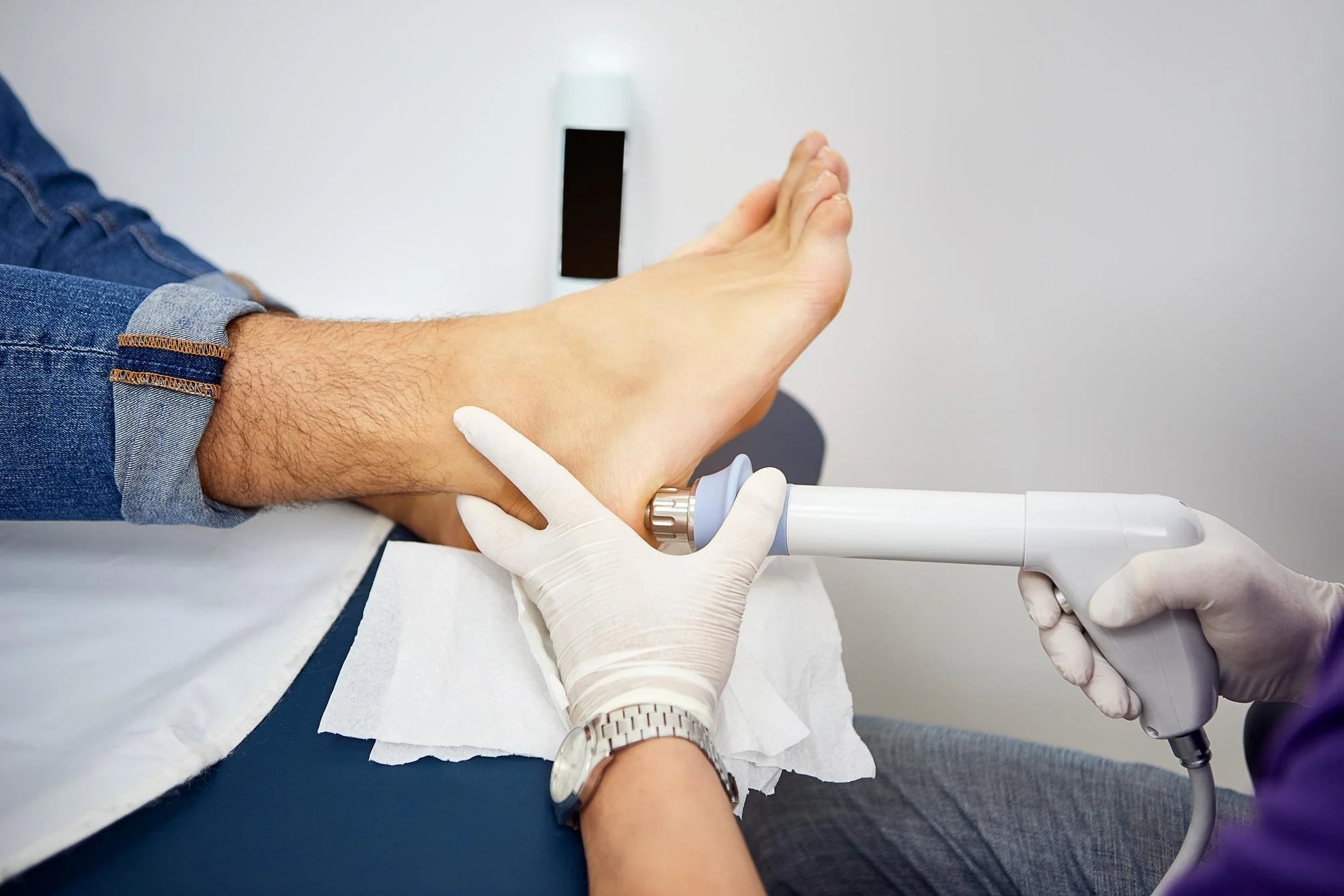 A person receiving a medical injection in the arm, with a healthcare professional administering it using a syringe. The person is lying on a medical bed with a white sheet, wearing a denim shirt pulled up to expose the arm.