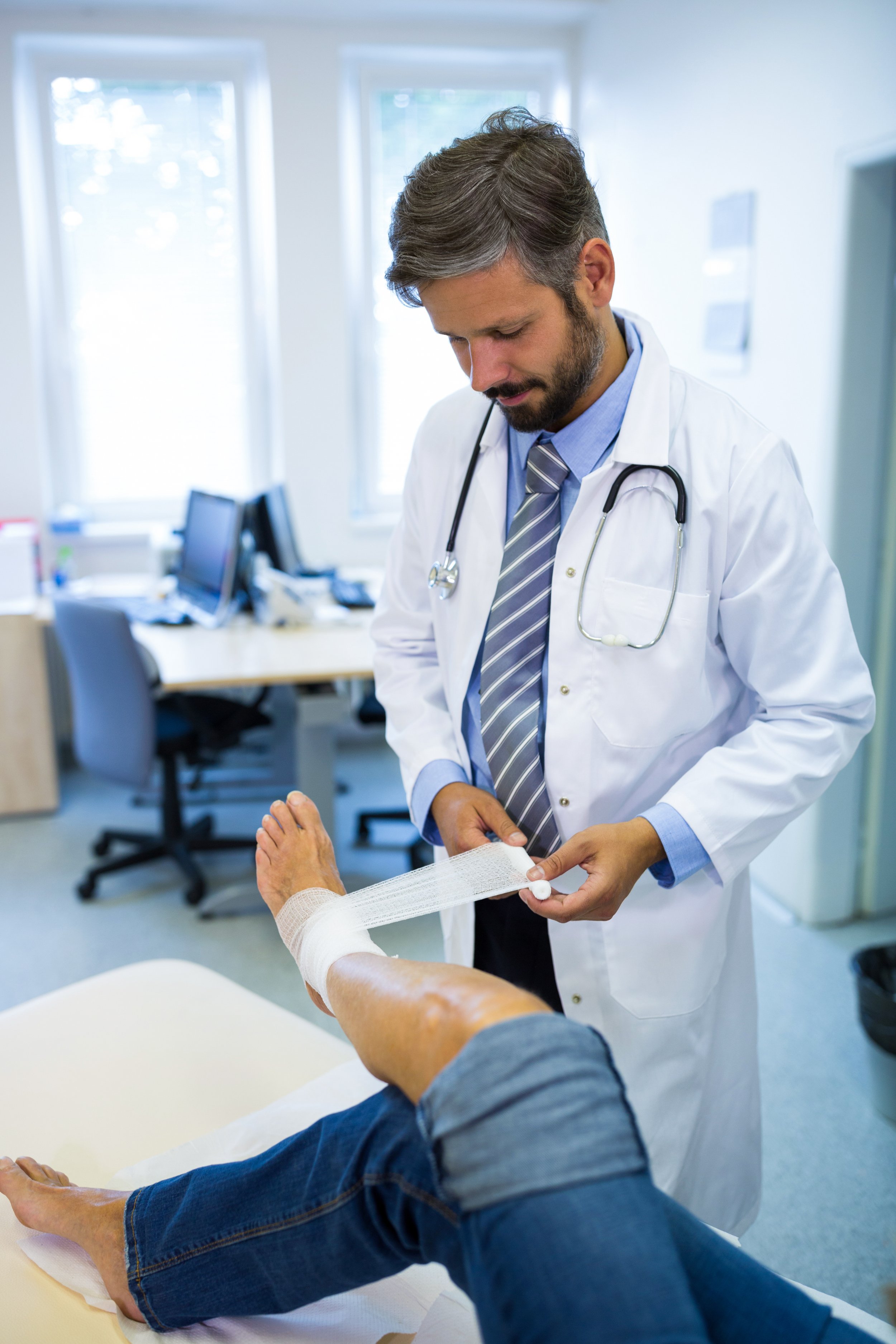 Doctor in a white coat with a stethoscope around his neck tending to a patient with a bandaged leg in a medical office.