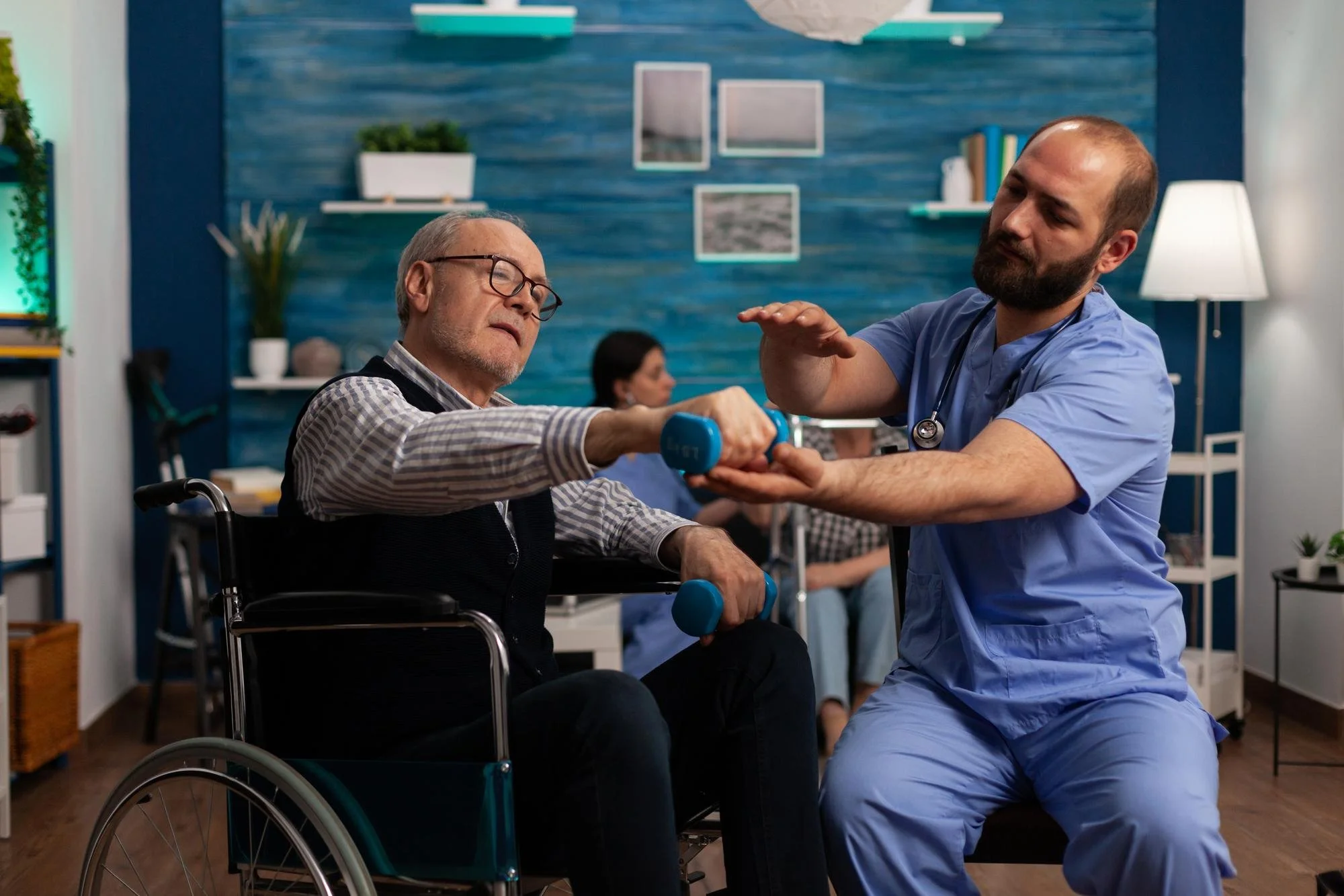 A healthcare professional in blue scrubs assisting an elderly man in a wheelchair with light blue dumbbells during physical therapy in a modern, well-lit clinic or hospital room.