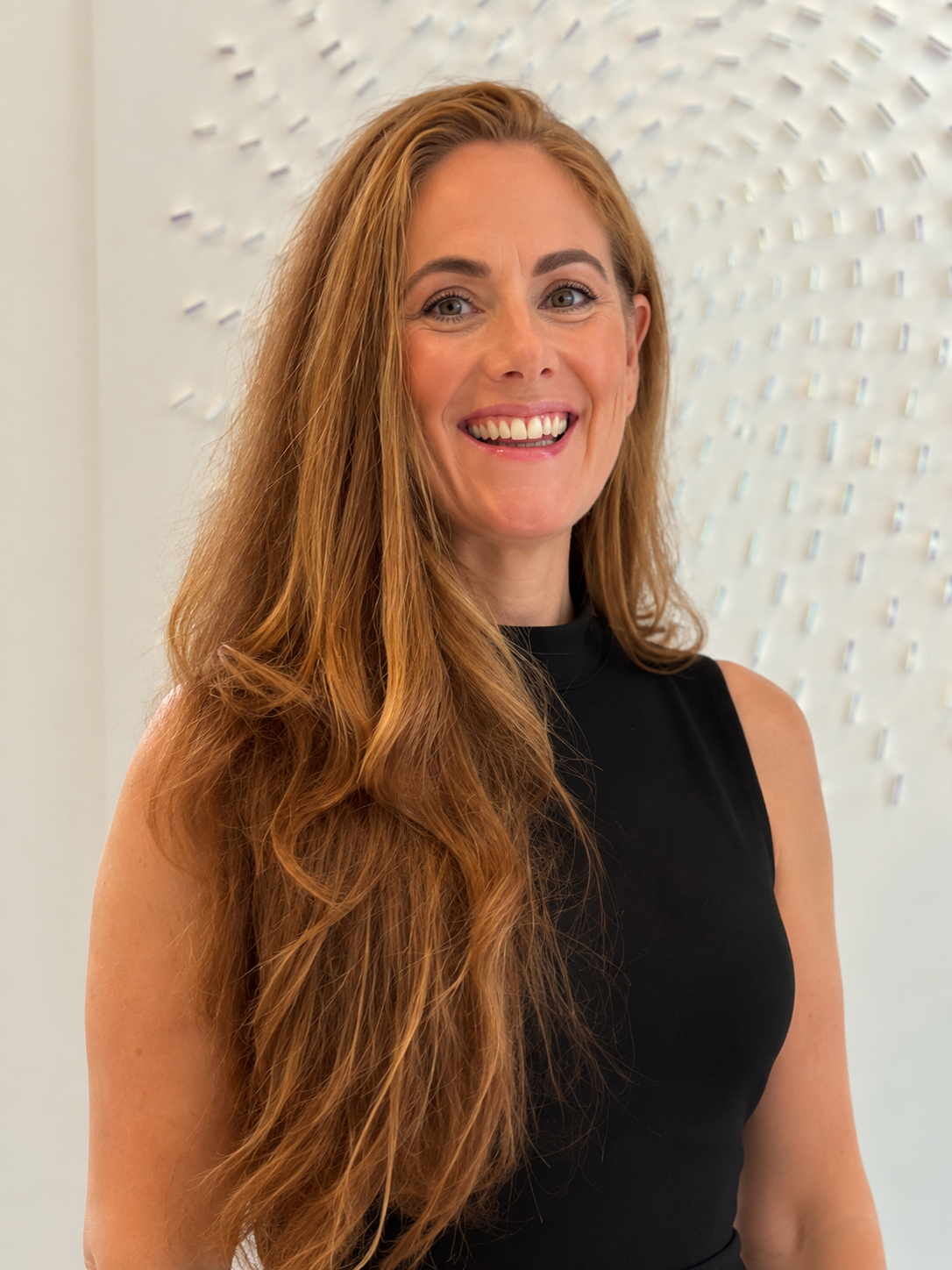 A woman with long red hair smiling, wearing a black sleeveless top, standing in front of a white textured background.
