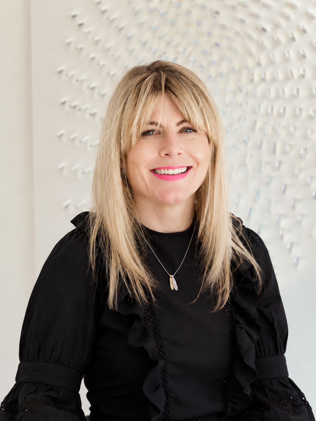 A woman with blonde hair smiling, wearing a black ruffled blouse and a silver necklace against a textured white wall background.