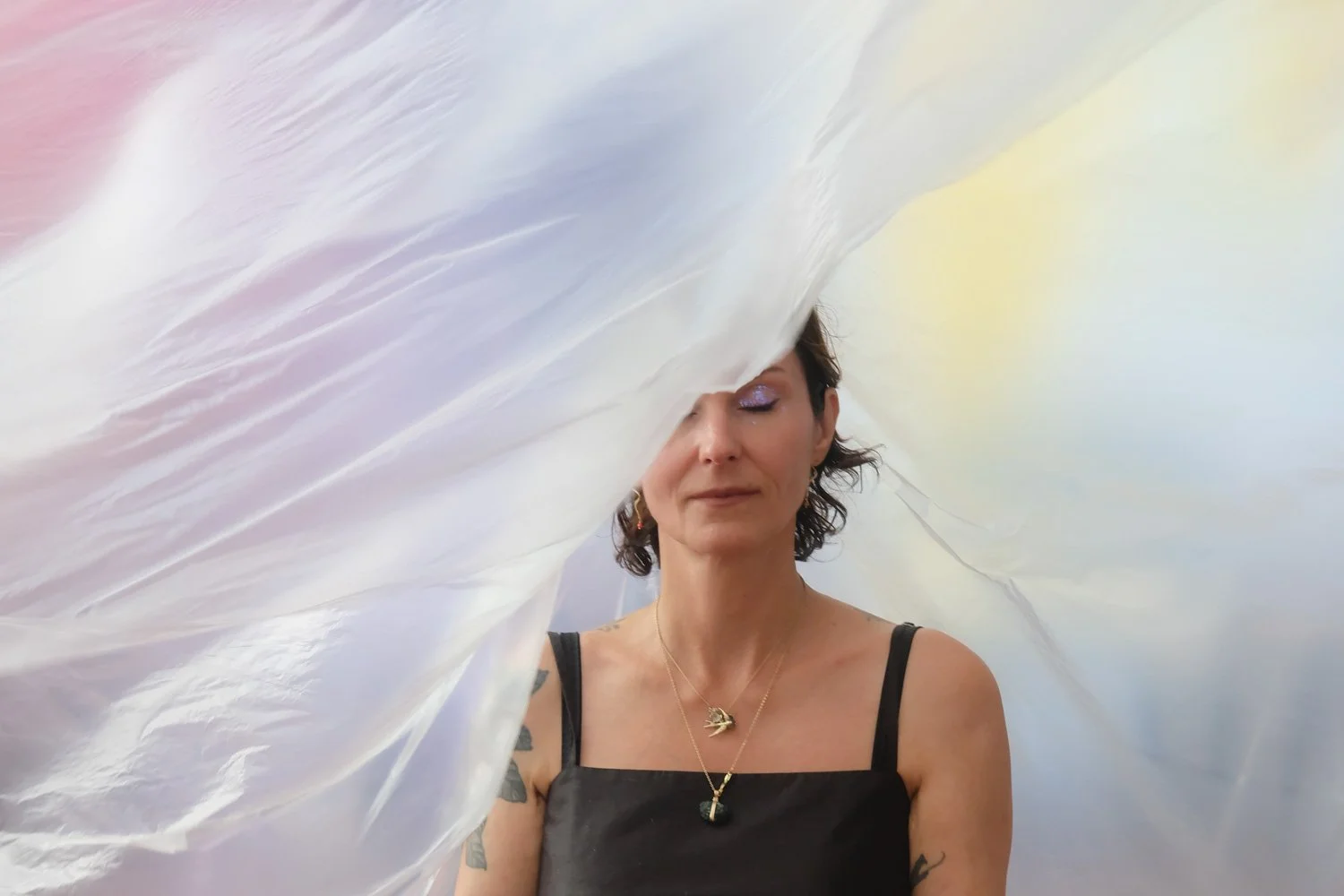 Vrouw met korte haar en tattoos op armen draagt een zwarte top en sieraden, met iriserende achtergrond, gedeeltelijk bedekt door een grote, kleurrijke, doorschijnende stof.