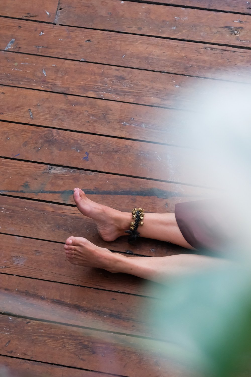 Voeten van een persoon met een sierlijke enkelband op een houten vloer, gedeeltelijk bedekt door een groene en witte vlek.