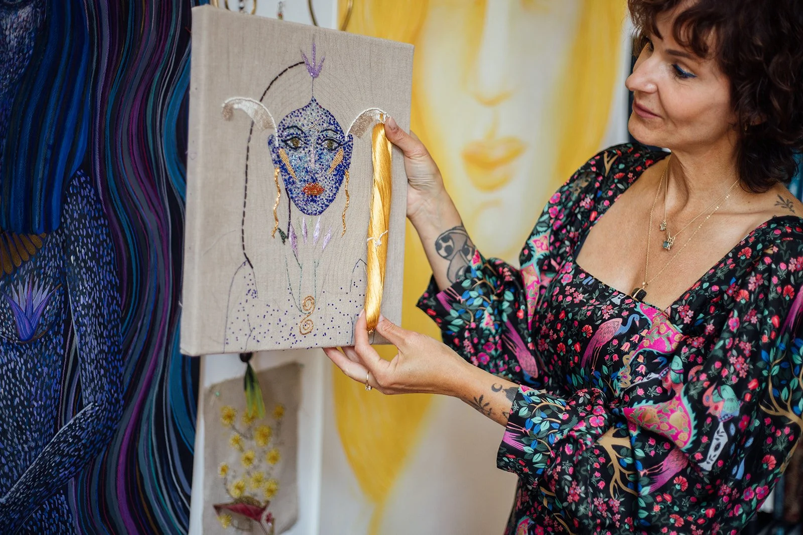 Een vrouw die een kunstwerk vasthoudt, dat een gezicht met sierlijke lijnen en strass-steentjes bevat, in een kunstgalerie met kleurrijke achtergrond.