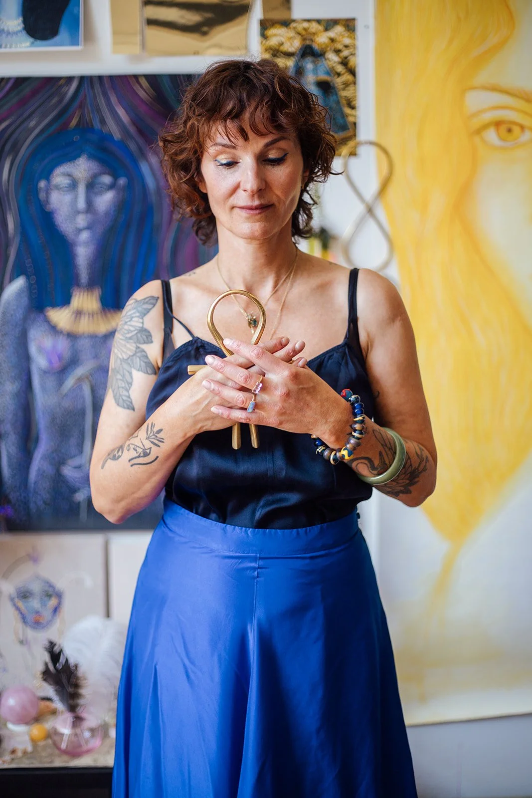 Vrouw met kort, krullend bruin haar, tattoos op armen, een ring en armbanden, houdt handen op haar hart en kijkt naar een hartvormige draagbare spiegel, standing in een kunstgalerie met kleurrijke kunstwerken op de achtergrond.