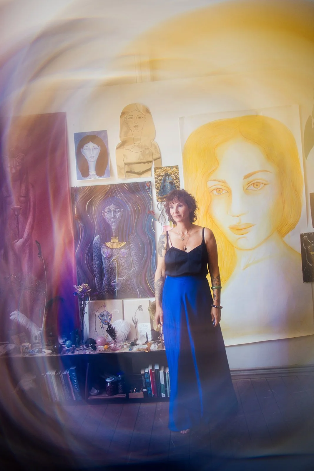 Een vrouw met tatoeages staat in een kamer vol kunstwerken en kunstobjecten.