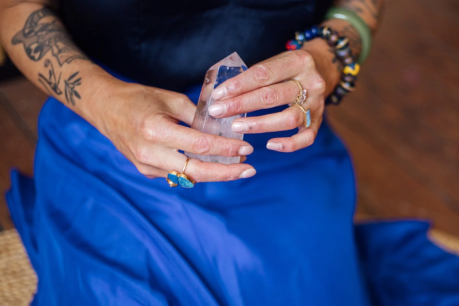 Close-up van een persoon die een kristallen beeld vasthoudt, met meerdere sieraden en tatoeages, zittend op een houten vloer, gekleed in blauwe broek en een zwart bovenstuk.