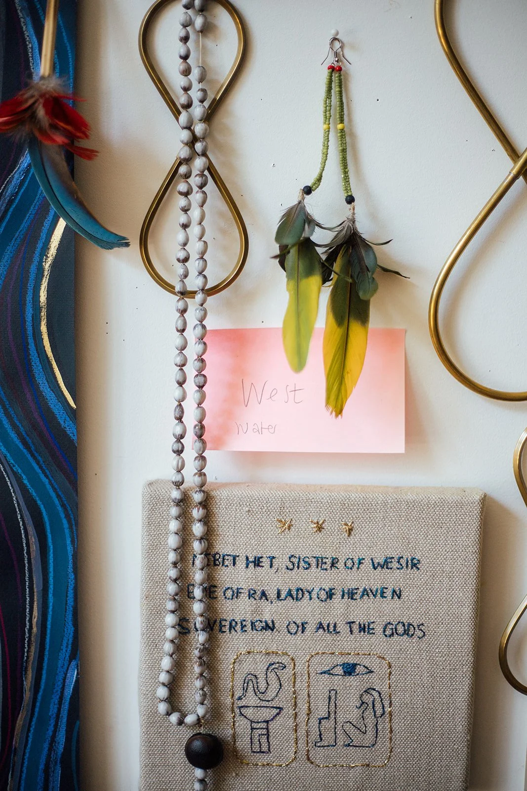 Een muur met diverse decoraties, waaronder een ketting van witte kralen, een hangertje met groene en gele veren, een bericht met 'West Water', en een bord met tekst en symbolen