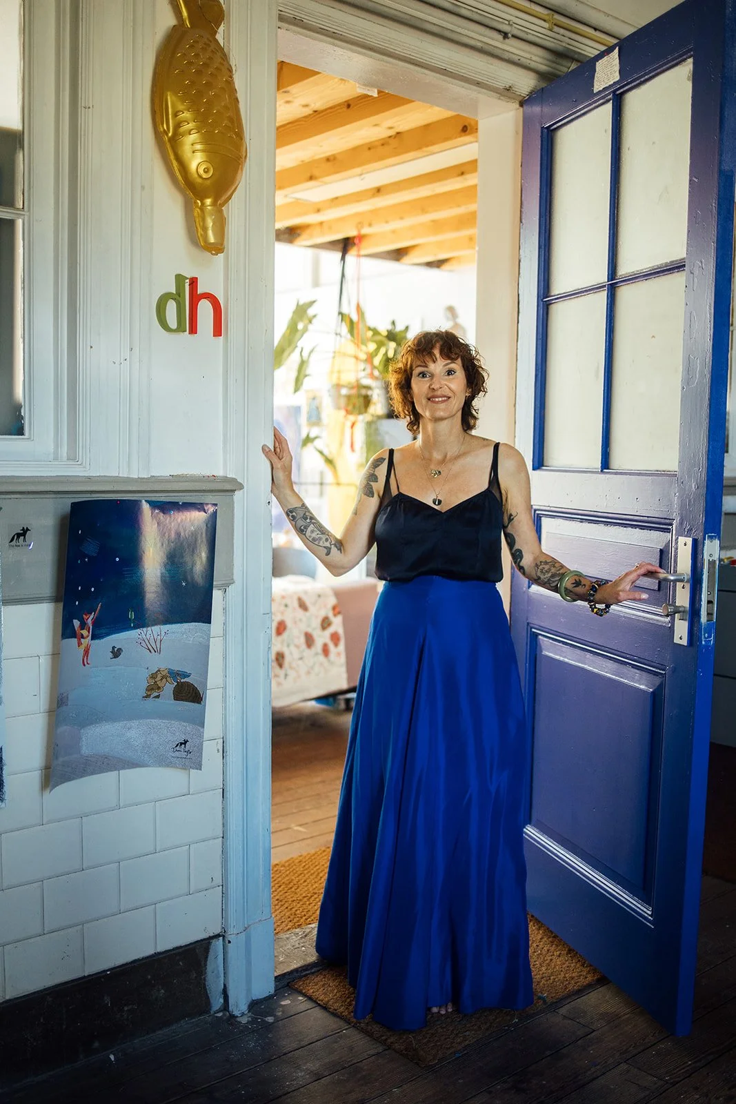 Vrouw met tattoos en blauwe rok staat in de deuropening van een huis, op een blauwe deur, met een open deur naar een binnenruimte met houten plafond en decoraties.