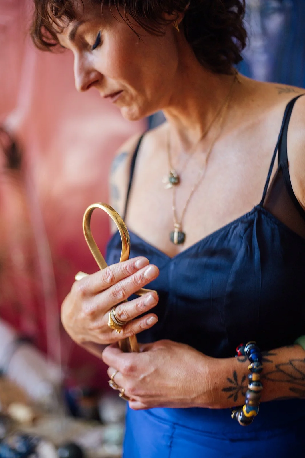 Vrouw die een gouden sieraad vasthoudt, met tattoos en accessoires, in een winkel met kleurrijke achtergrond.