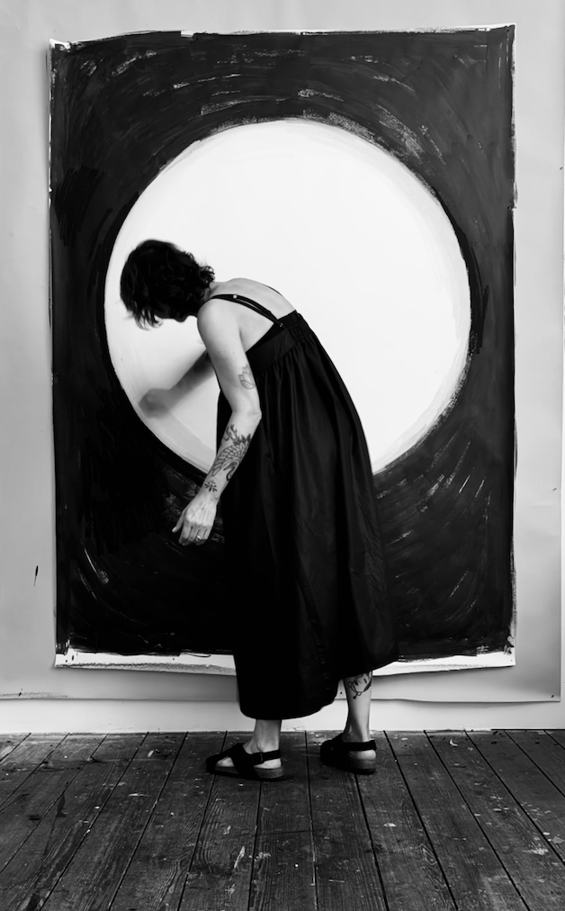 Vrouw met tattoos en zwarte jurk die een grote, abstracte zwart-witte schilderij aan het maken is.