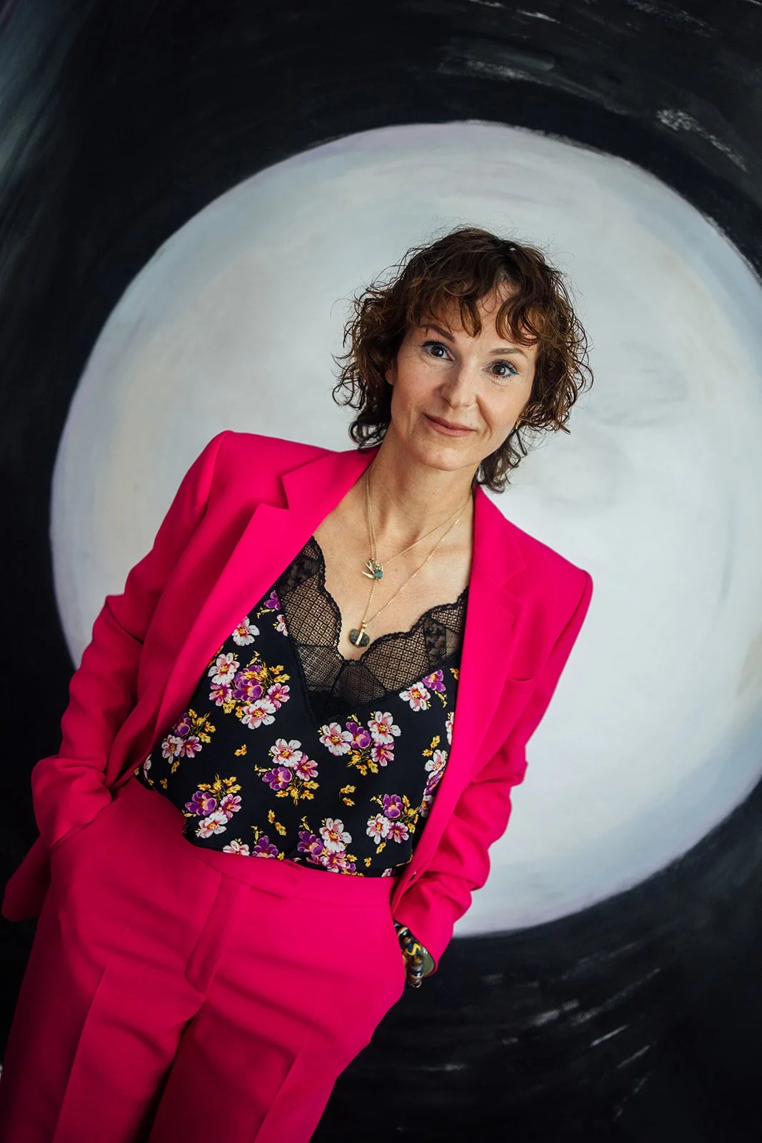 Portret van een vrouw met kort, golvend bruin haar, gekleed in een fuchsia roze broek en blazer, met een zwarte top met bloemenpatroon en kanten details, staand voor een muur met een cirkelvormig zwart-wit kunstwerk.