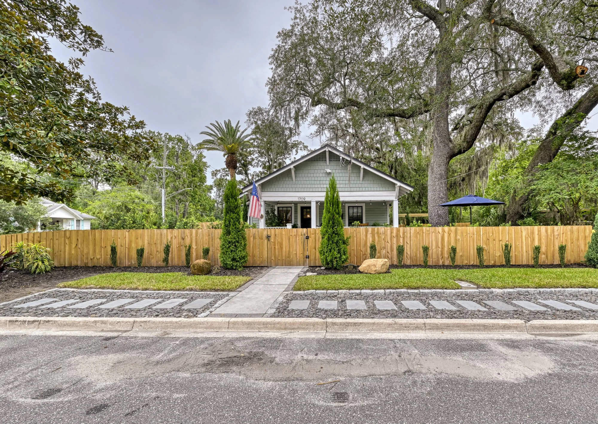 A house with a gray exterior, front porch, and the number 1709 on it, surrounded by a wooden fence, with a large tree and palm tree behind it, and a yard with rocks and plants in front.