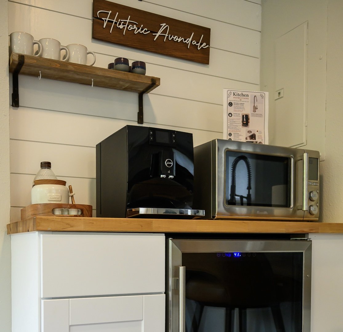 Coffee machine, microwave, and mini fridge on a wooden countertop in a kitchen with white shiplap walls, wooden shelves with mugs and cups, and a sign that says 'Historic Avondale'.