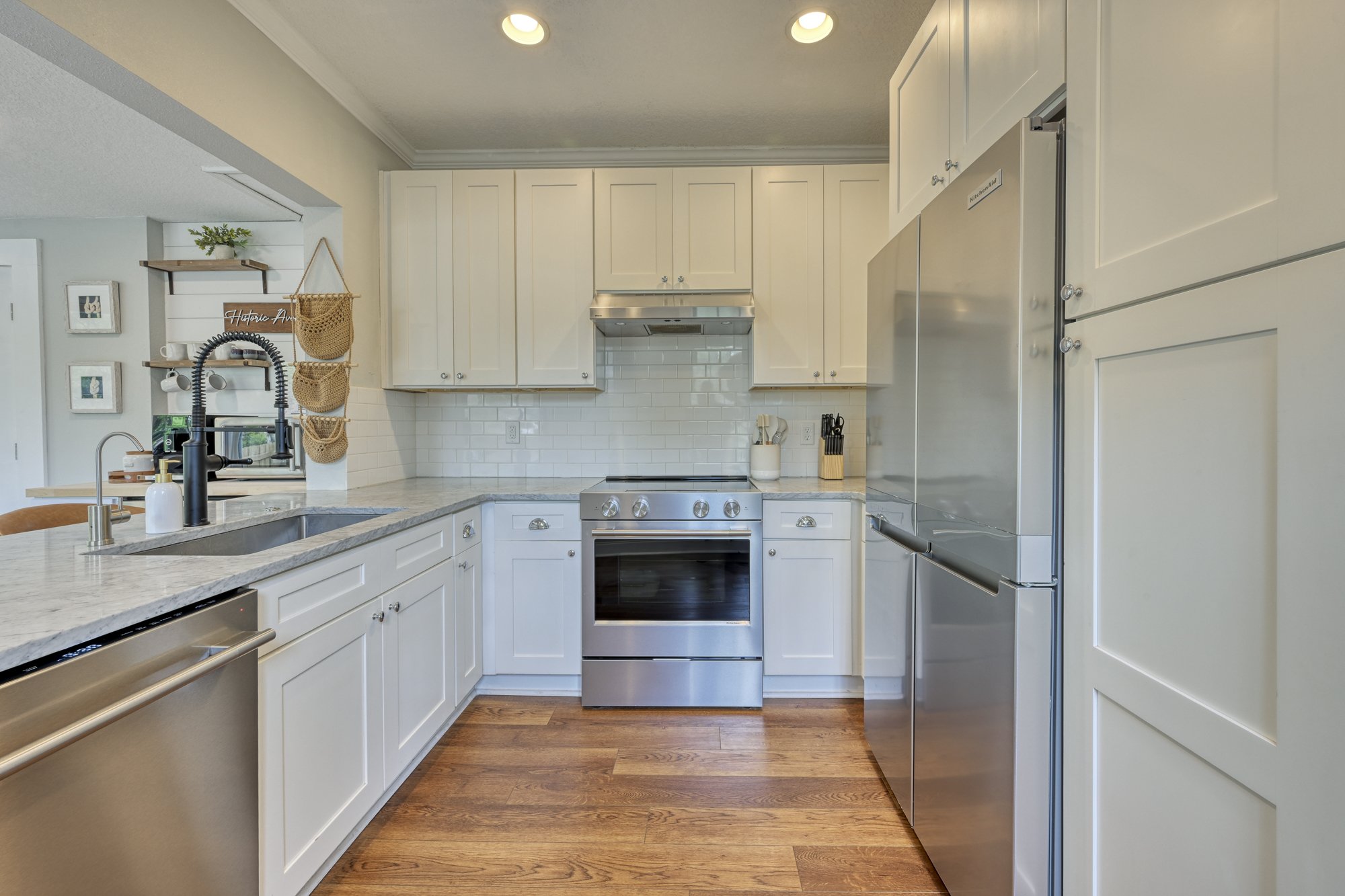 Kitchen with white cabinets, stainless steel appliances, wood flooring, and a marble countertop, with various kitchen accessories and decor.