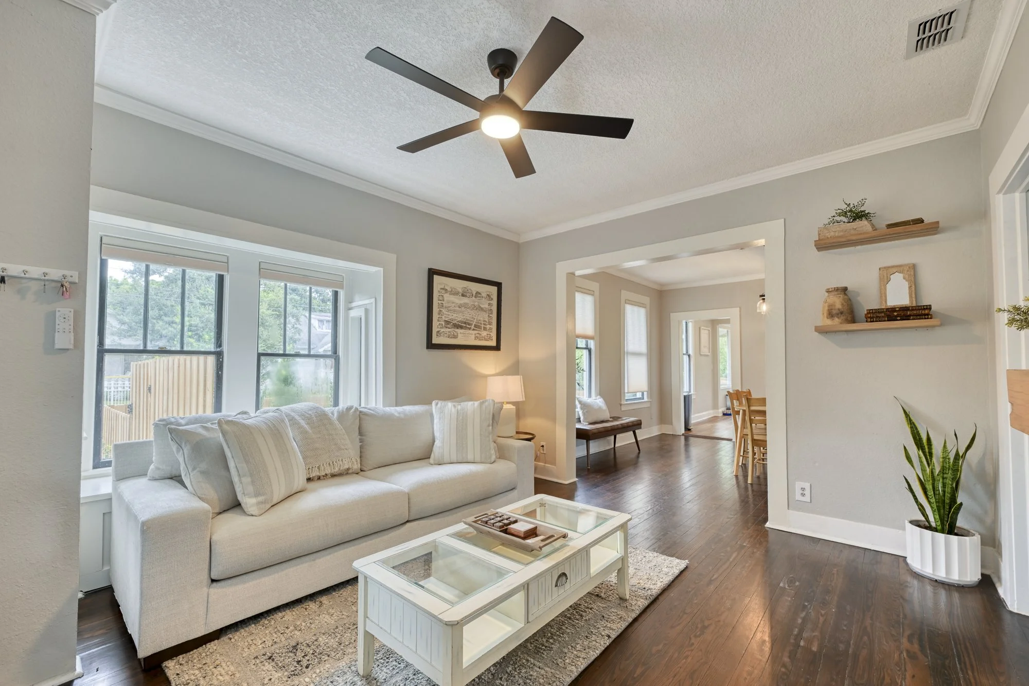 Living room with white sofa, coffee table, wooden floors, walls painted light gray, ceiling fan with three blades, natural light from large windows, shelving with decorative items, and houseplants.