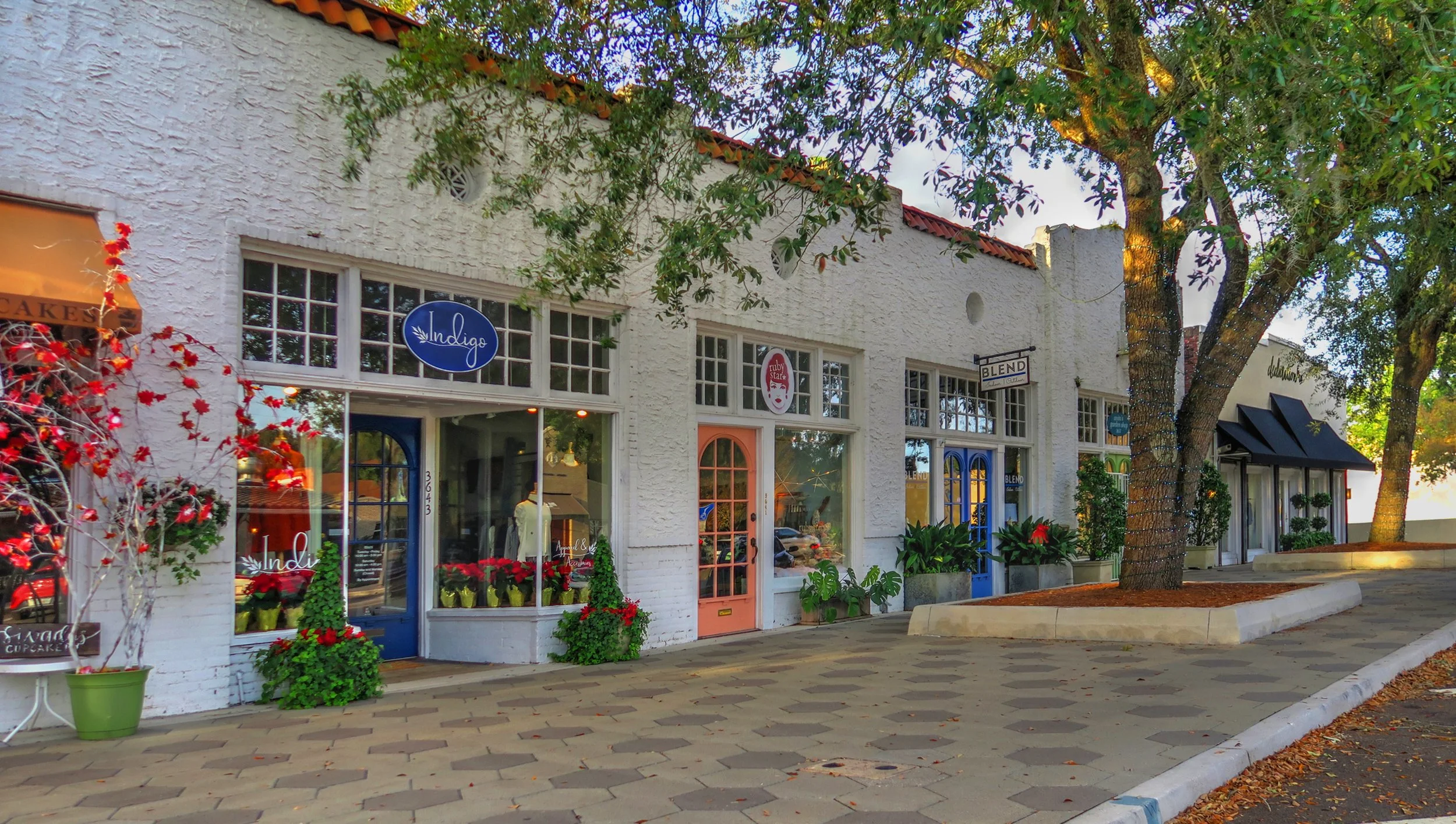 A row of storefronts in a small shopping district, including Indie and Blend stores, with trees and outdoor decorations, and a stone sidewalk in front.