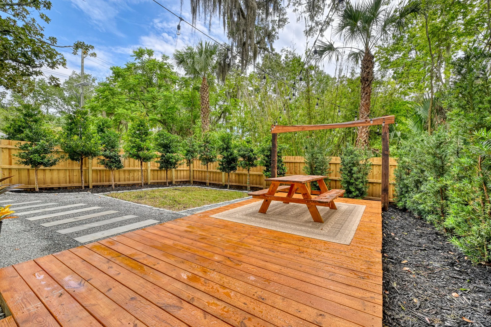 A backyard outdoor area featuring a wooden deck with a picnic table and a shaded pergola, surrounded by lush green trees and bushes, with a gravel pathway to the left and string lights hanging between trees.