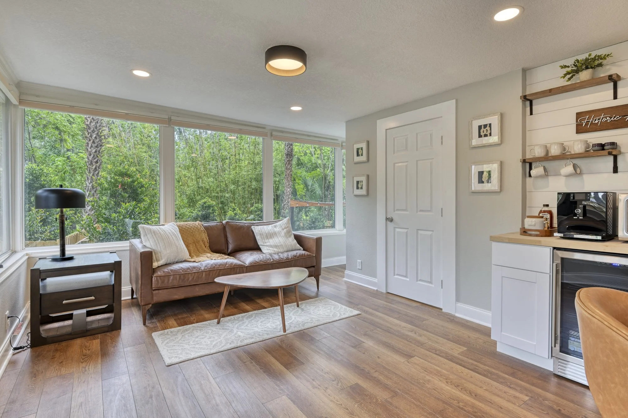Living room with large window showing trees outside, brown leather sofa with pillows, black table lamp, small coffee table, white cabinets, and shelves with kitchen items, hardwood flooring, and ceiling lights.