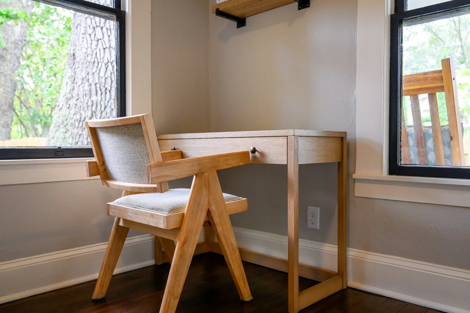 Corner of a room with a wooden desk and matching chair near large windows showing trees outside, and a wooden shelf mounted on the wall.
