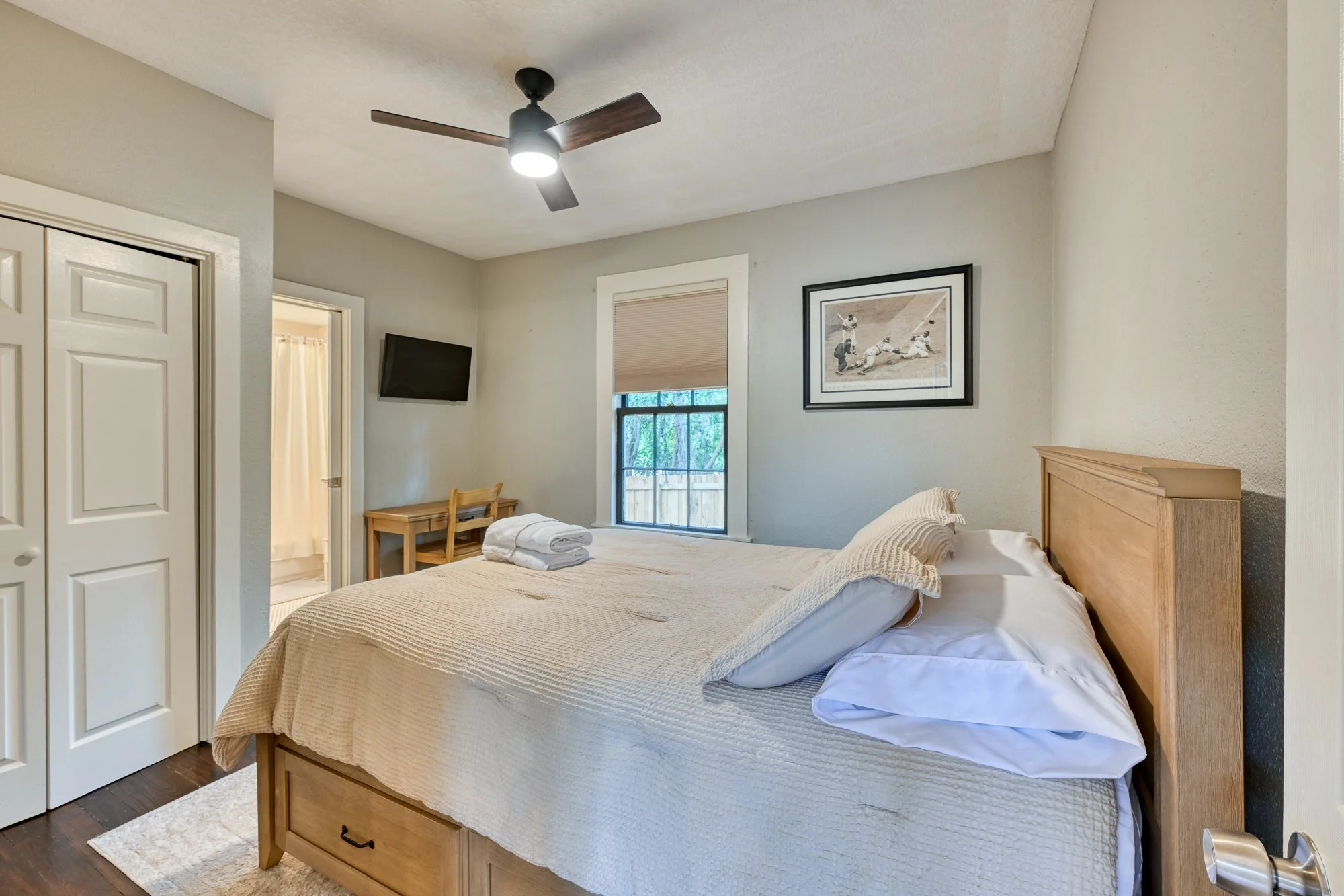 A bedroom with a bed, a window with blinds, a picture frame on the wall, a ceiling fan, a small table with a chair, and a flat-screen TV mounted on the wall.