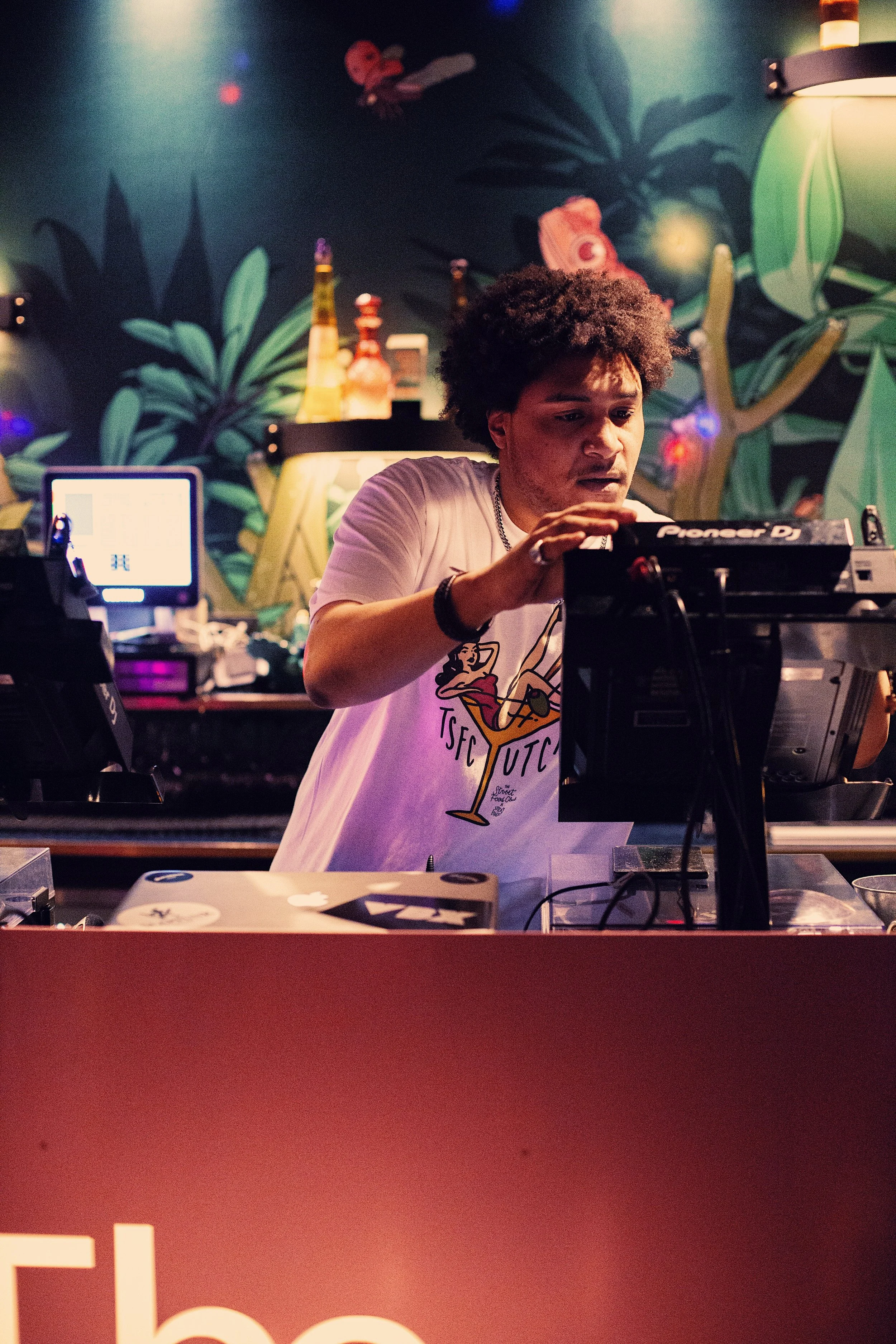 Een persoon met afro haar die DJ-setup bedient in een ruimte met een groene en blauwe muur met botanische patronen, verlicht door kleurrijk licht.