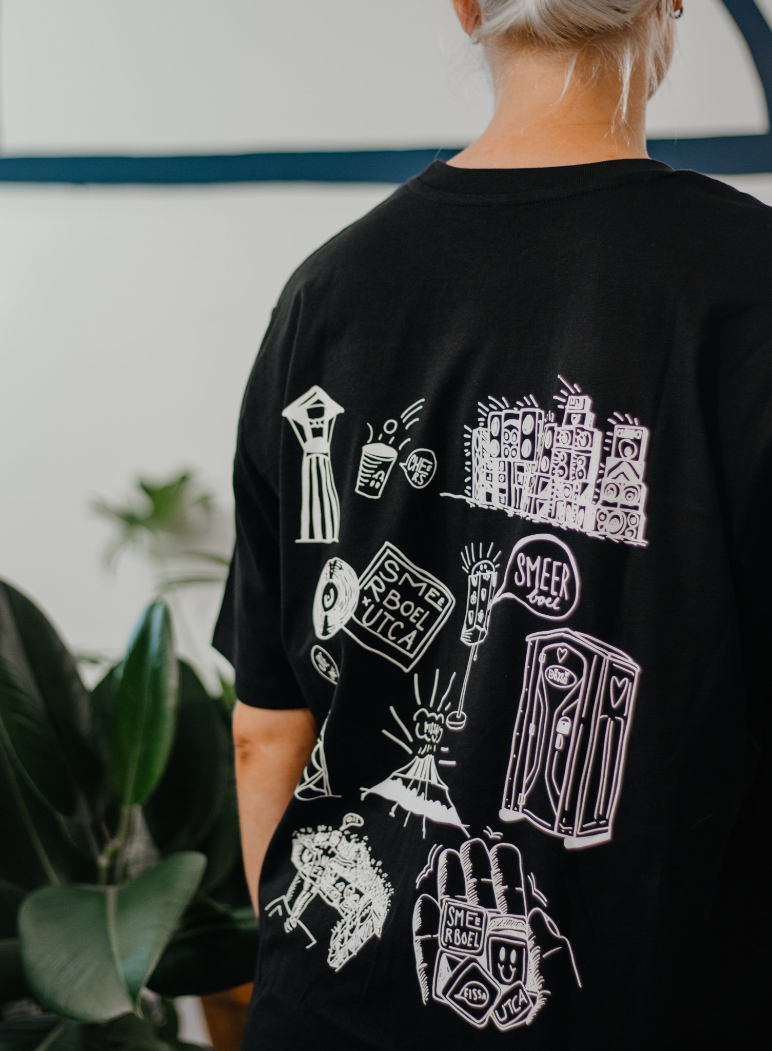 Close-up van een persoon met een zwart T-shirt met witte illustraties van verschillende voorwerpen en woorden, te zien vanaf de achterkant.