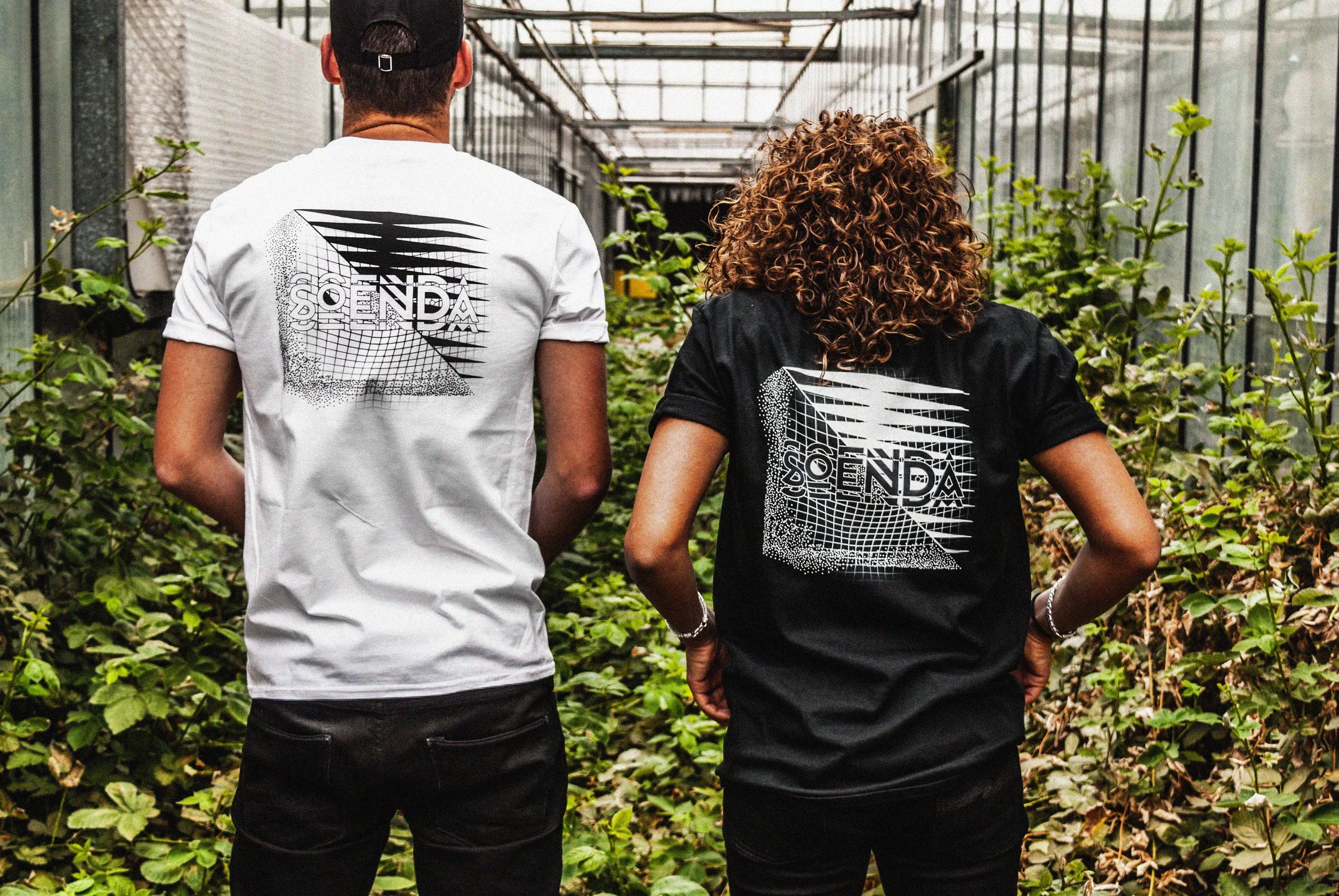 SOENDA festival t-shirt design door Utca's Finest, Twee mensen lopen door een kas met groene planten, ze dragen T-shirts met hetzelfde geometrische ontwerp en tekst 'SOENDA' op de achterkant.