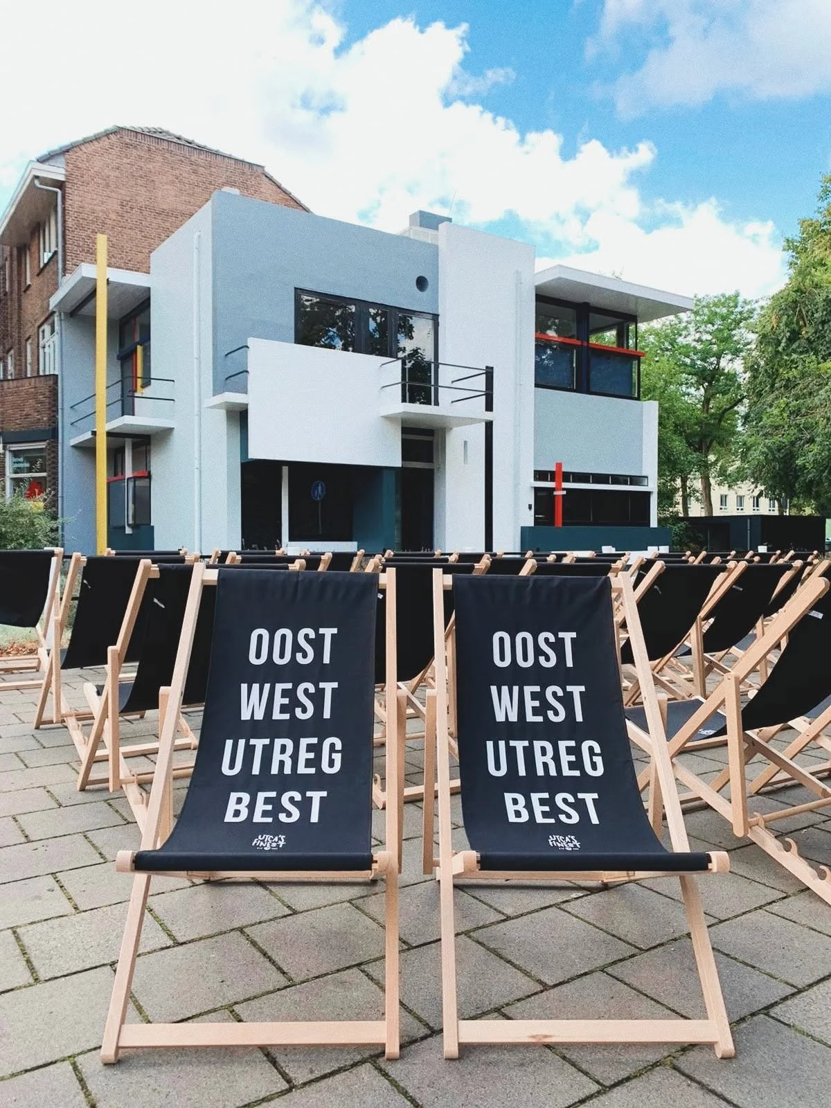 Strandstoelen met tekst 'OOST WEST UTREG BEST' van Utca's Finest voor een moderne witte gebouw genoemd het Rietveld Schröderhuis met grote ramen, in de buitenlucht op een zonnige dag.
