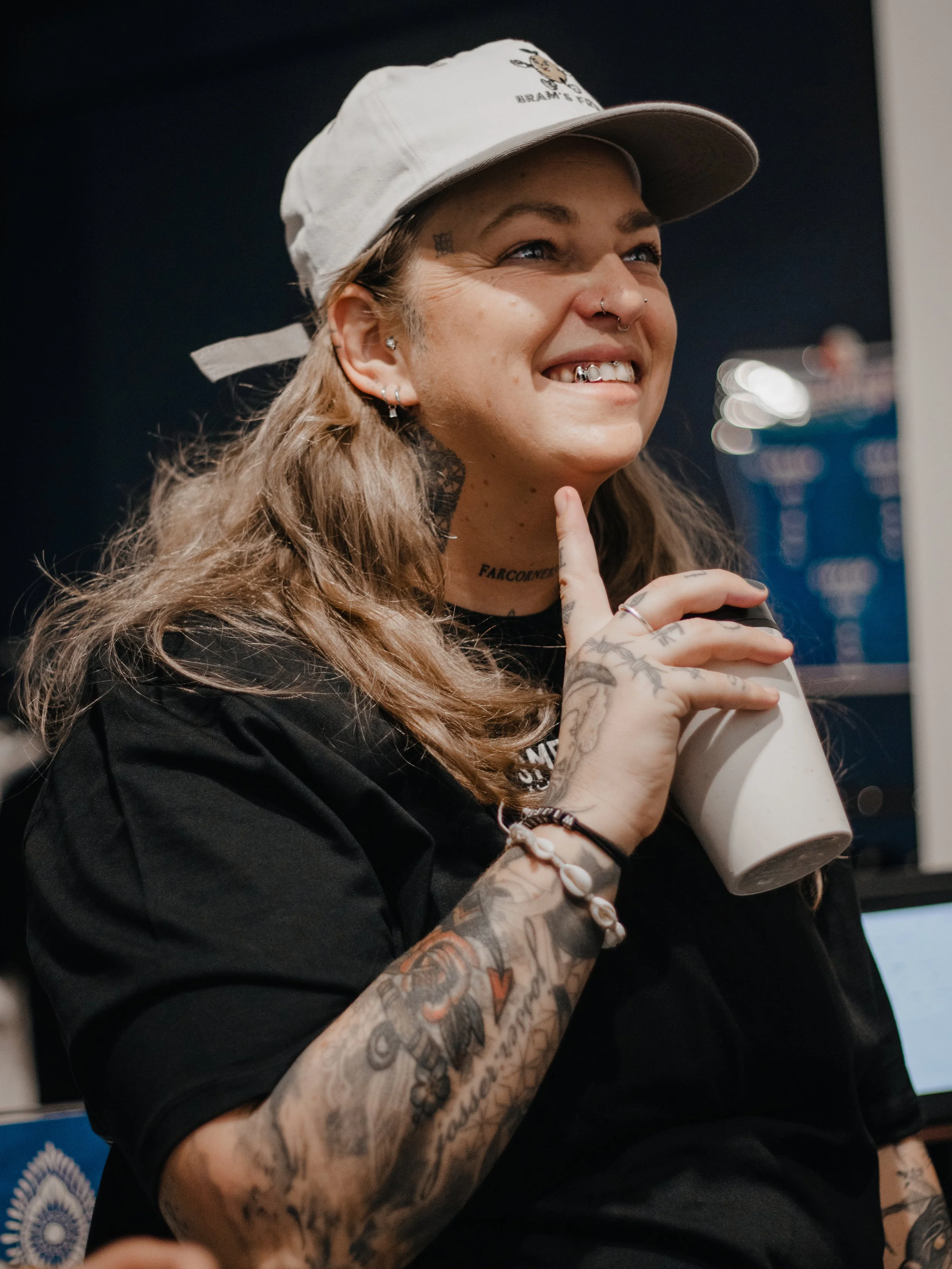Close-up van een glimlachende vrouw met tattoos en piercings, die een witte koptelefoon vasthoudt, in een donkere achtergrond.