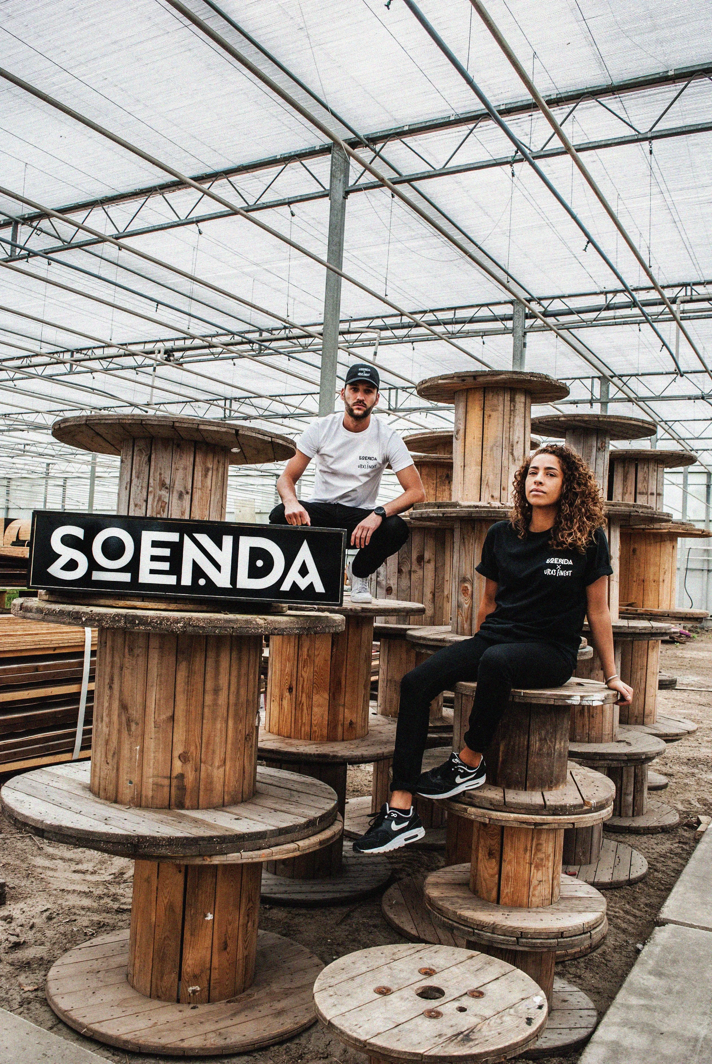 Twee jonge mensen poseren tussen grote houten spools in een kas of magazijn, met een bord dat 'SOENDA' zegt en een kleinere tekst eronder.